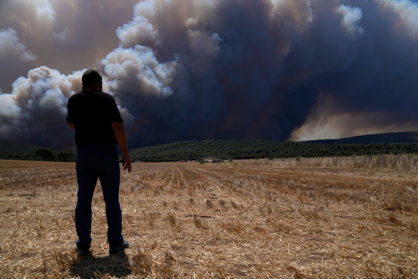 Ein Mann steht auf einem Feld, während im Hintergrund der Rauch eines Feuers in der Nähe von Athen zu sehen ist.