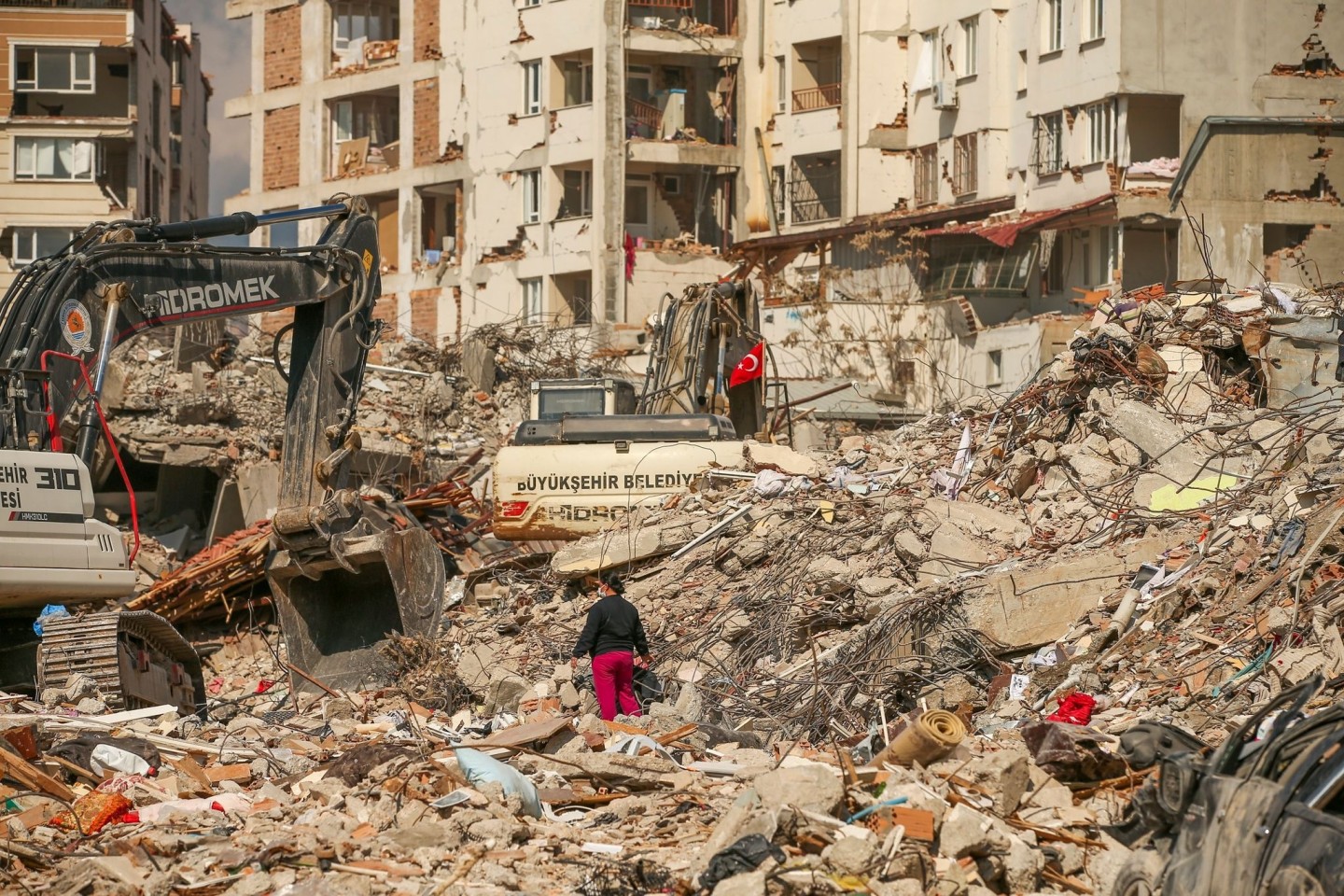 Ein Mann steht in Samandag in der Türkei zwischen Trümmern, während Bagger die Überreste eines zerstörten Gebäudes abtransportieren. Nach dem katastrophalen Beben am 6.2. kommt es in d...