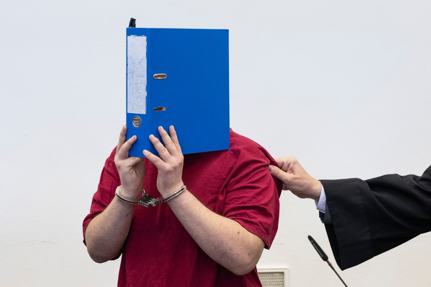 Der Angeklagte steht hinter der Anklagebank und verdeckt sein Gesicht hinter einem Aktenordner.