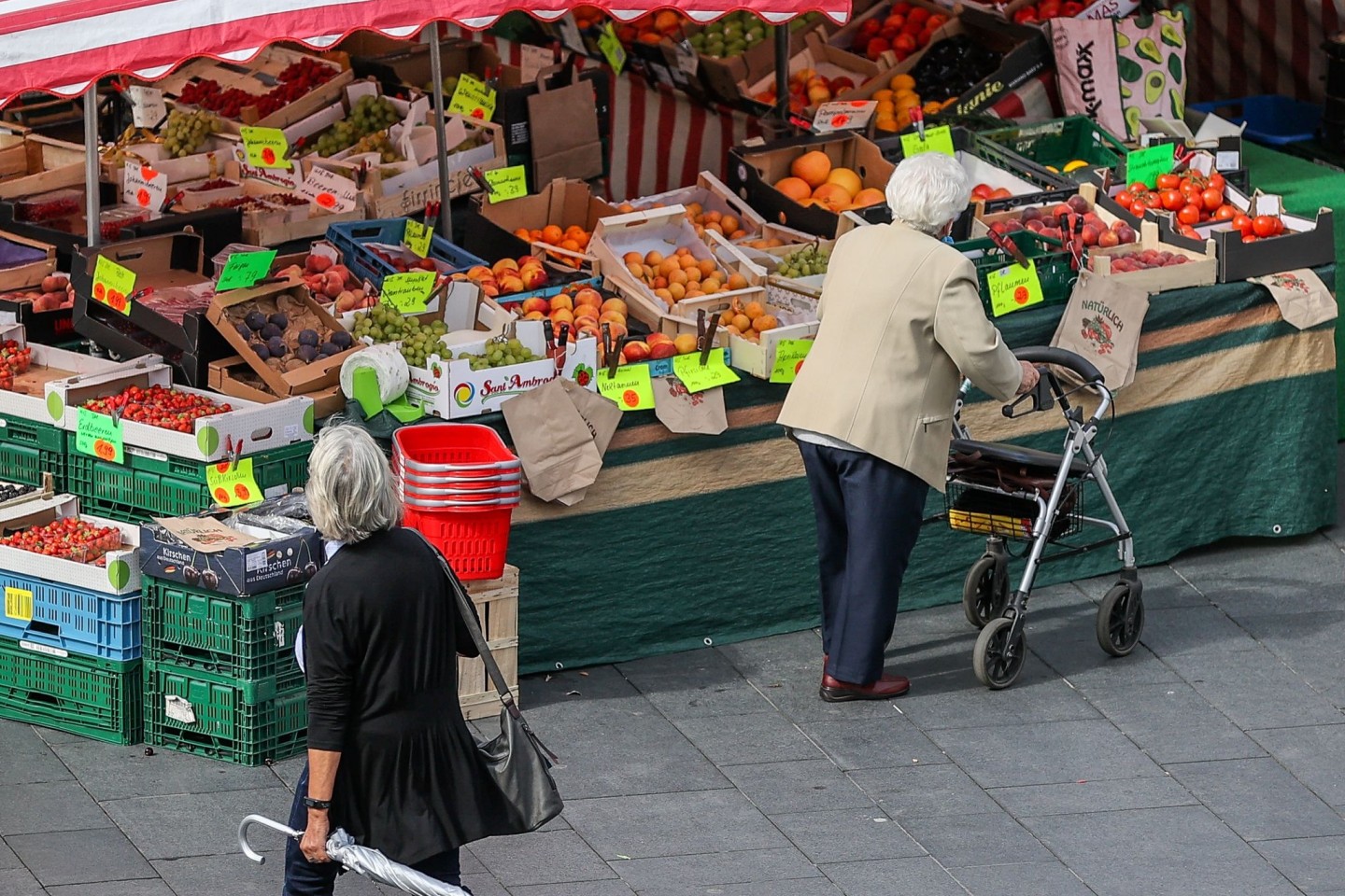 Eine Seniorin steht, gestützt auf ihren Rollator, vor einem Obst- und Gemüsestand auf einem Wochenmarkt.