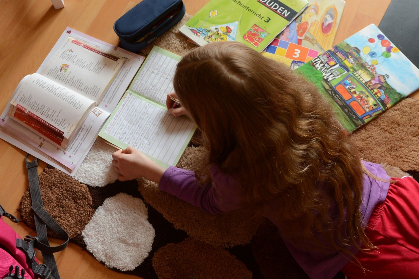 Ein Mädchen liegt auf einem Teppich in seinem Kinderzimmer und erledigt seine Hausaufgaben im Fach Deutsch.